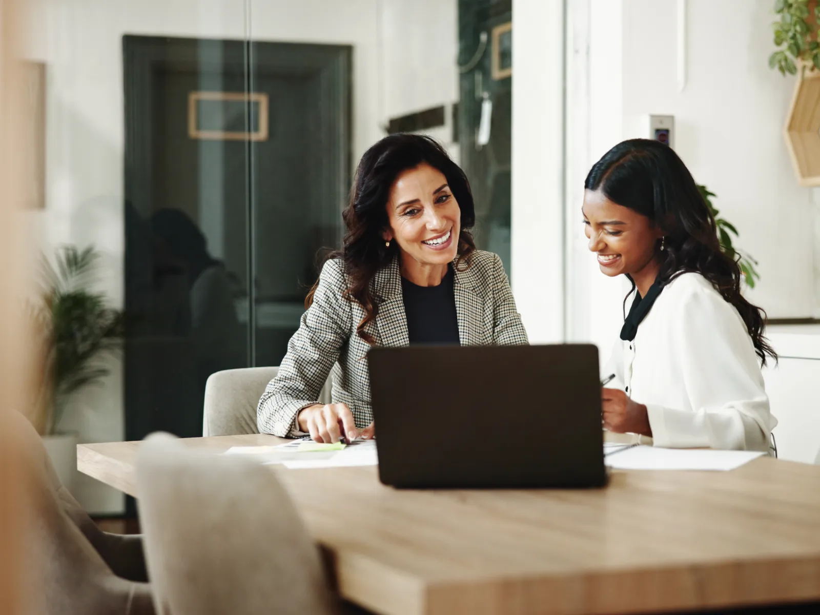 Two professional women smiling and discussing work over a laptop in a modern office setting.