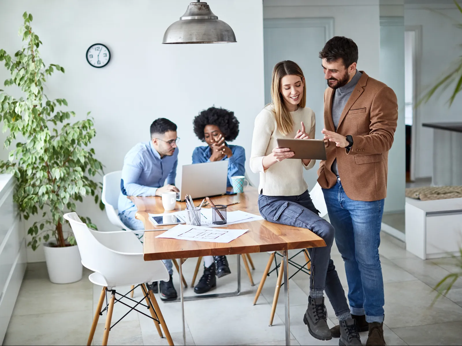 Four young professionals collaborating in a modern office with laptops and tablet around a wooden table.