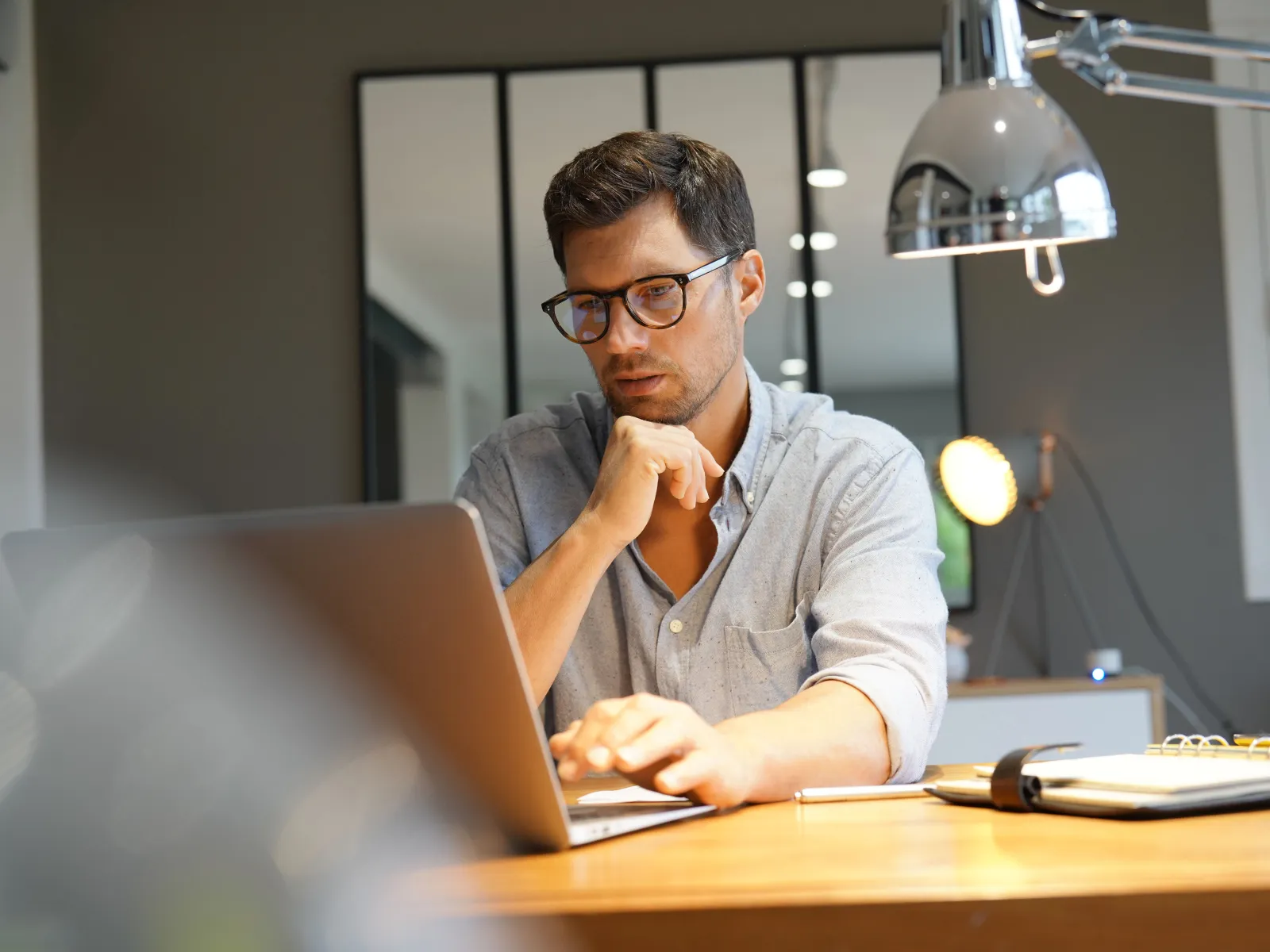 Man in glasses working thoughtfully on a laptop at a wooden desk in a modern home office.