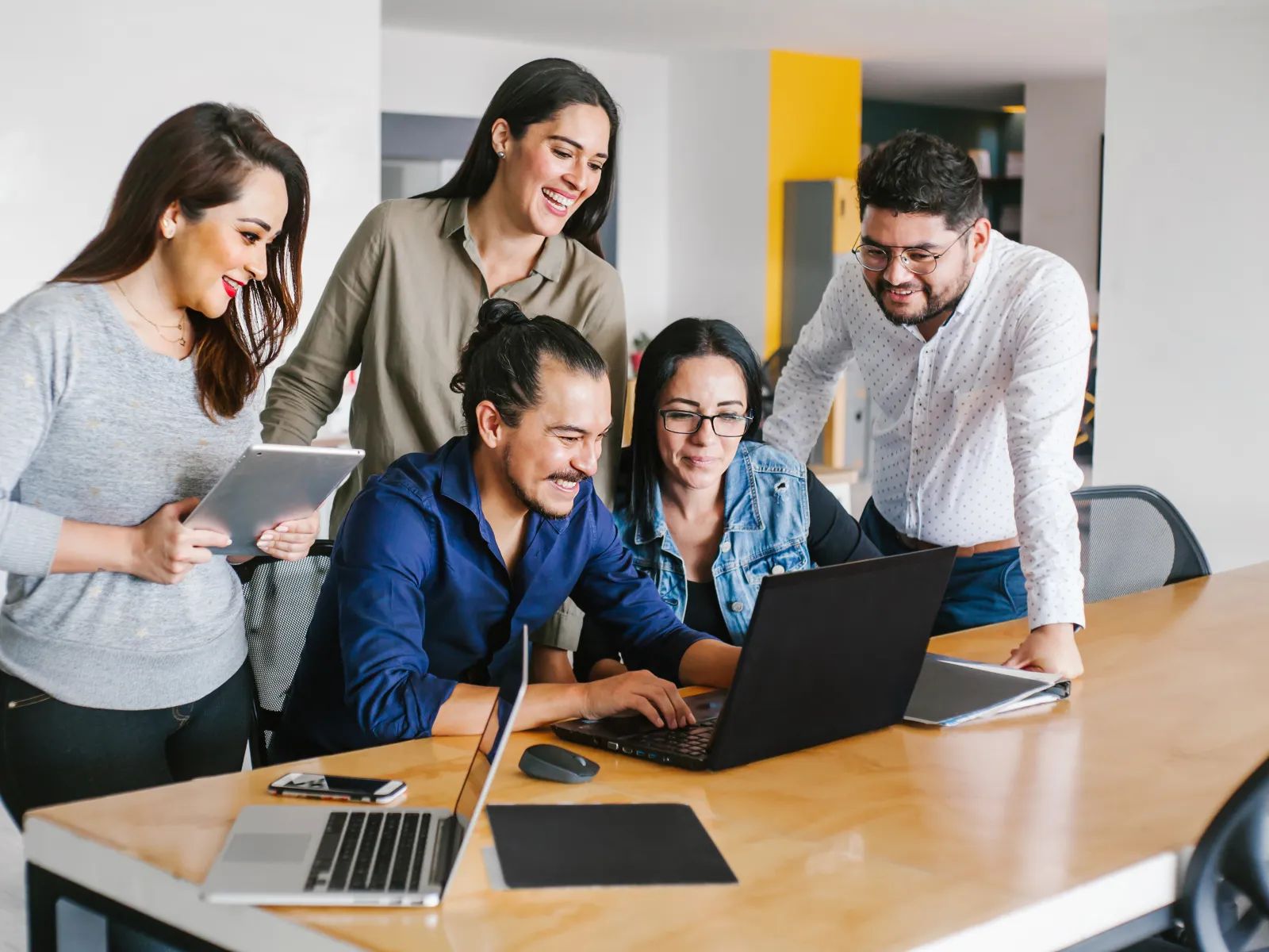 Five young professionals collaborating at a desk with laptops and a tablet in a modern office space