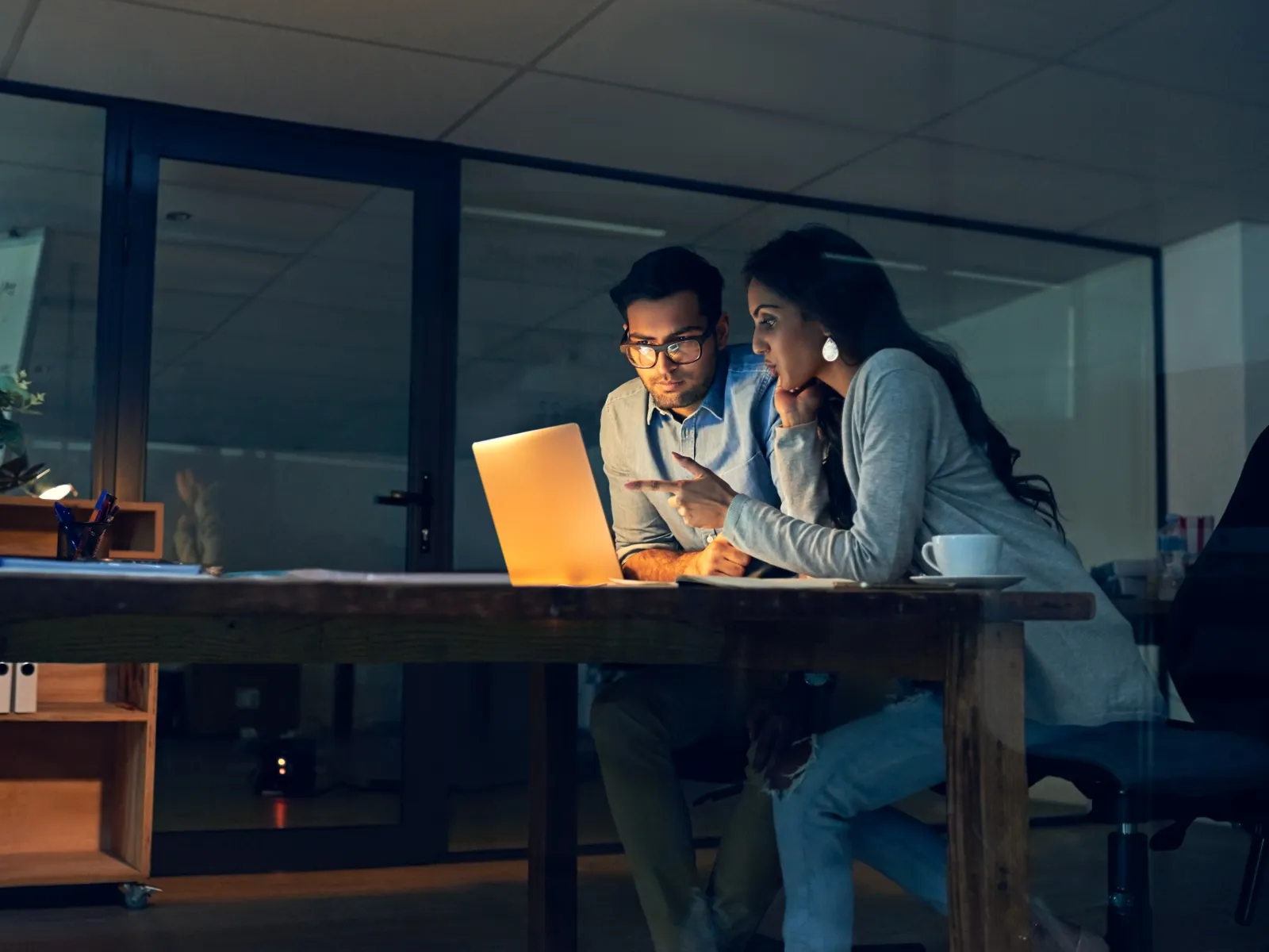 Two young professionals working late at night collaborating over a glowing laptop in an office meeting room.