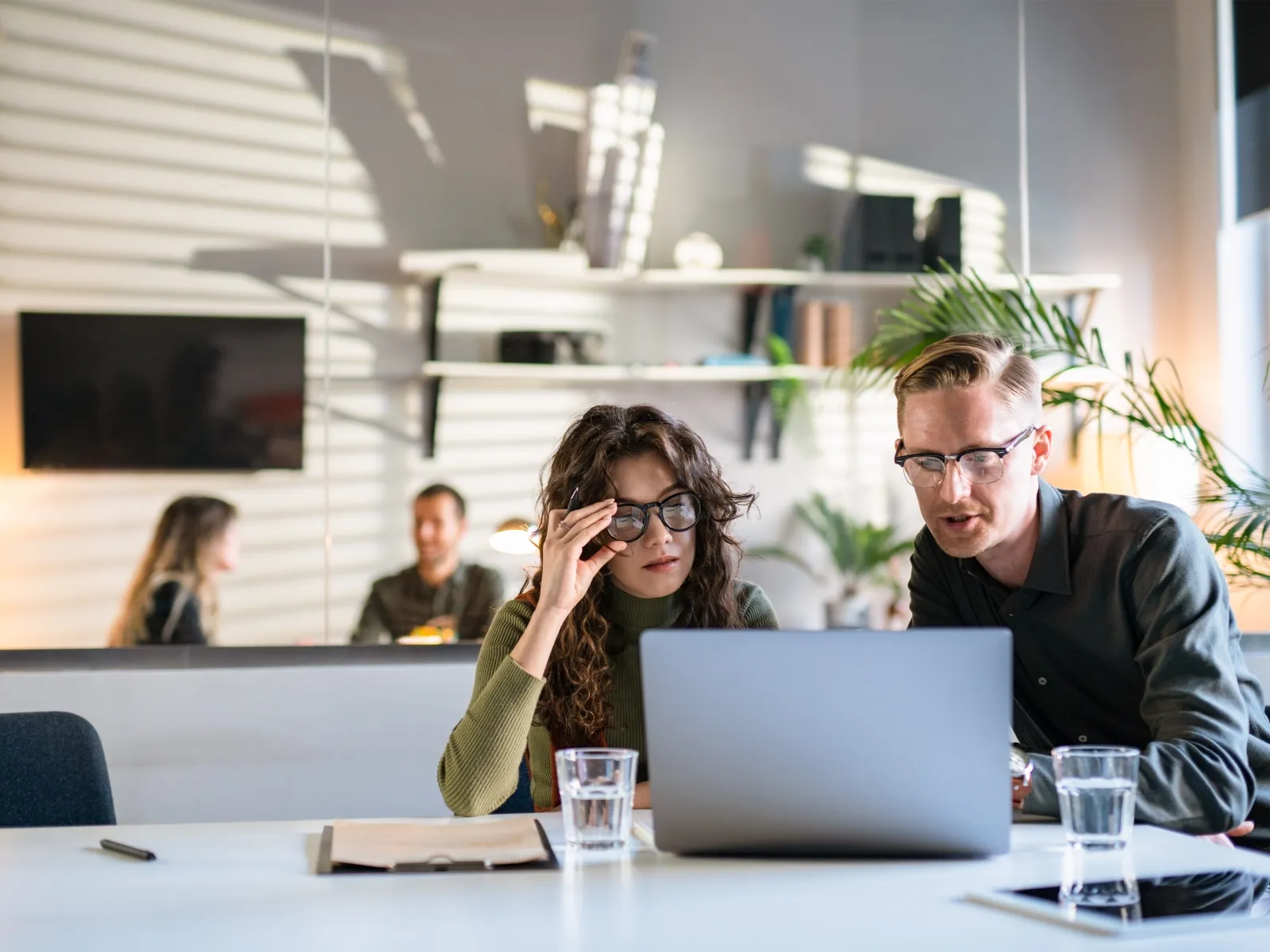 Two colleagues wearing glasses collaborate on a laptop in a modern office with plants and natural light.