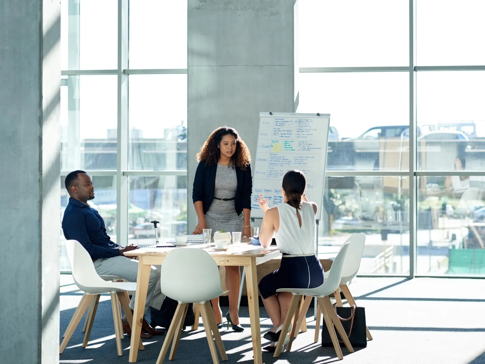 Three professionals having a meeting around a table with a whiteboard in a spacious modern office with large windows.
