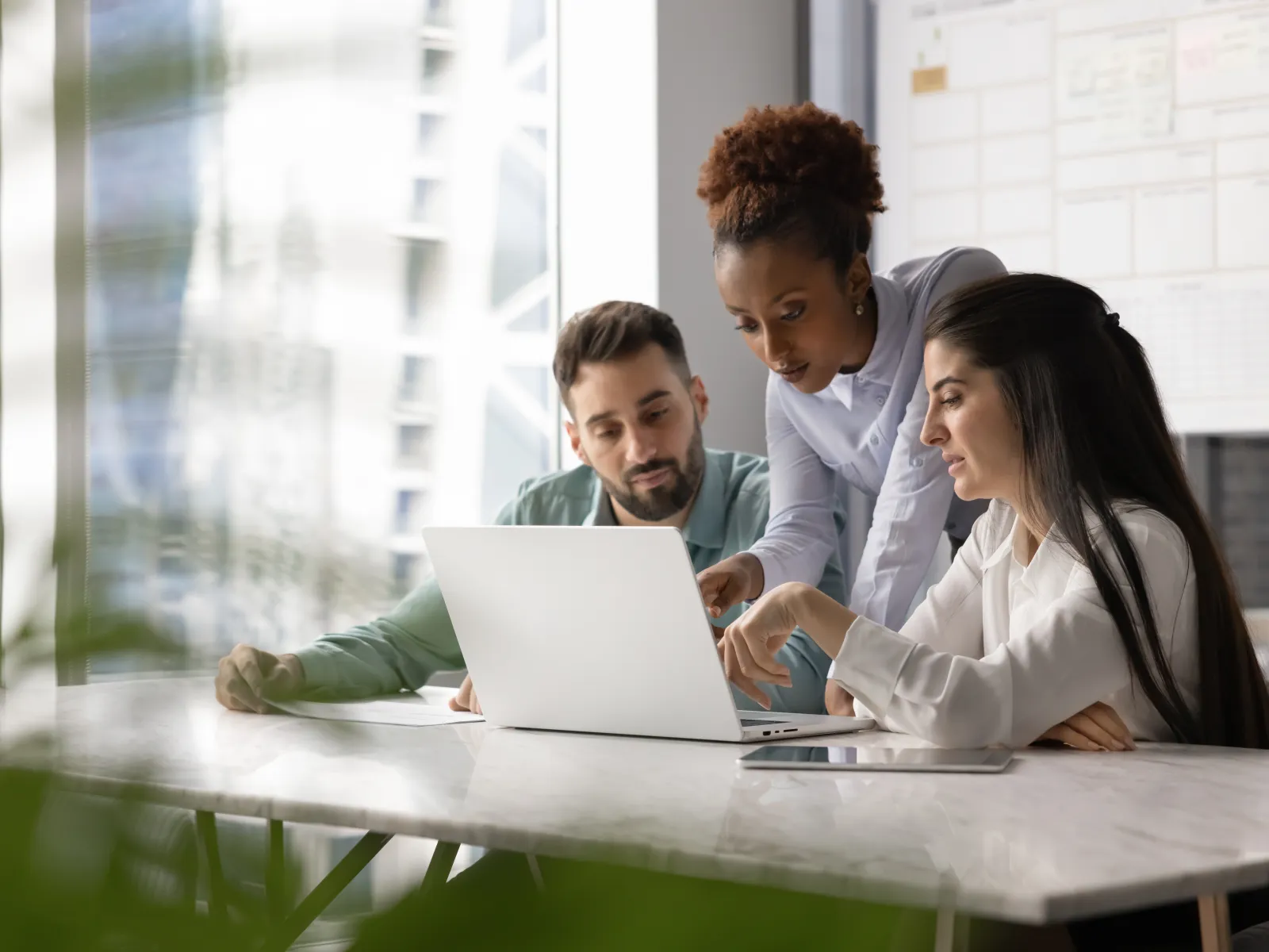 Three coworkers collaborate over a laptop at a modern office table by a large window during a meeting.