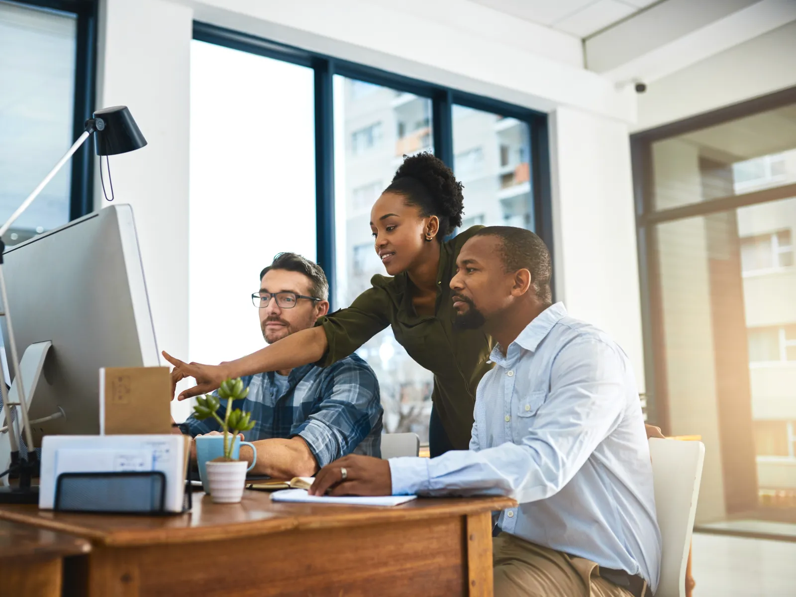 Diverse team collaborating on computer work in modern office with large windows and natural light.
