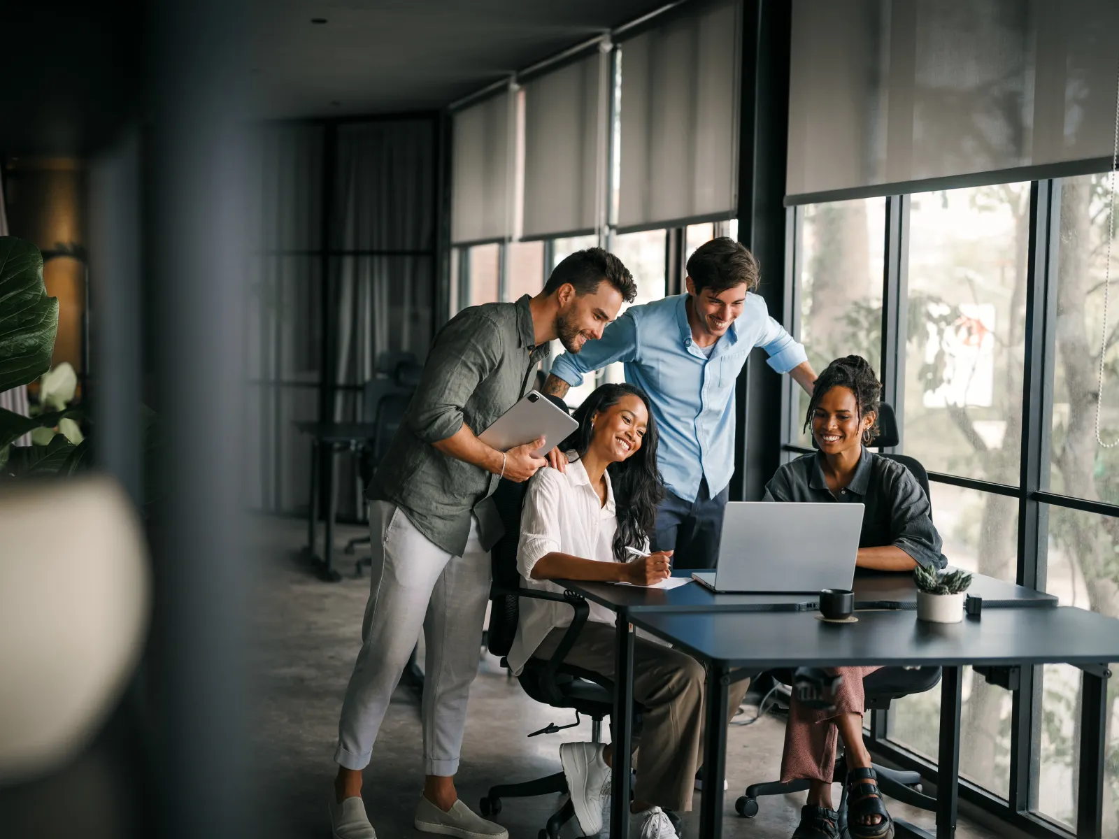 Four diverse coworkers smiling and collaborating around a laptop in a modern office with large windows.