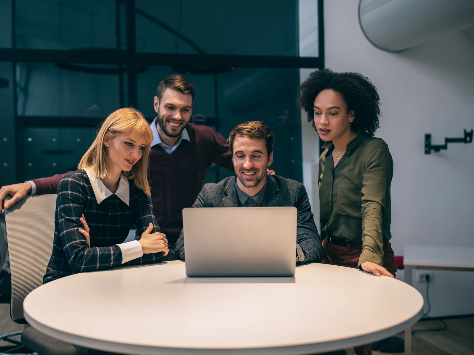 Four diverse colleagues gathered around a laptop at a round table in an office, collaborating and smiling.
