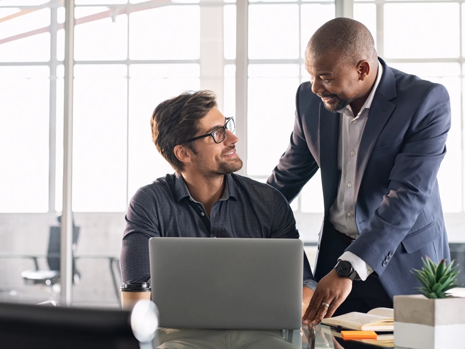 Two business colleagues discussing work with a laptop in a modern office setting near a window.