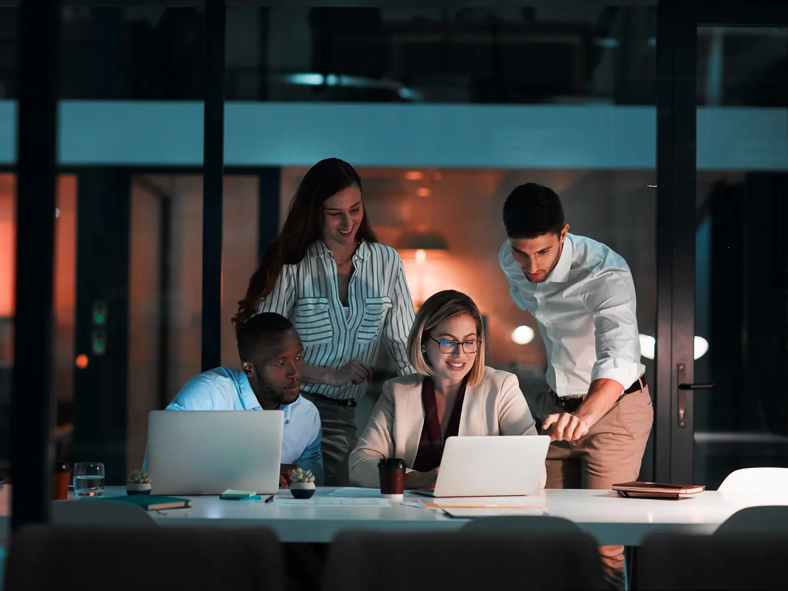 Diverse team collaborating in modern office at night around laptops discussing project ideas together
