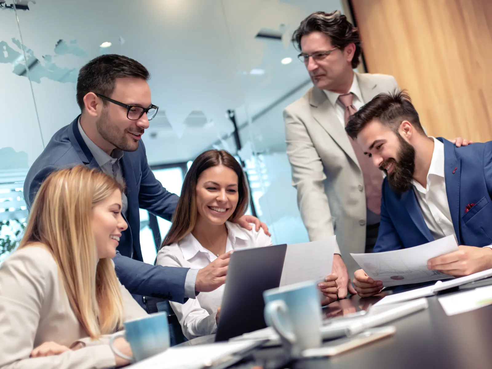 Group of five diverse business professionals collaborating over documents and laptop in a modern office meeting.