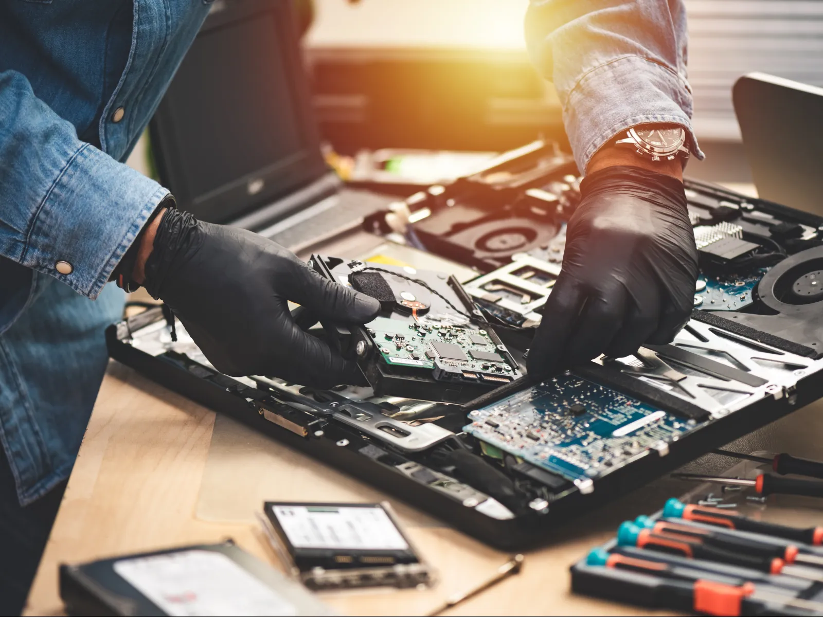 Technician wearing gloves repairing a laptop motherboard with tools on a wooden desk under sunlight.