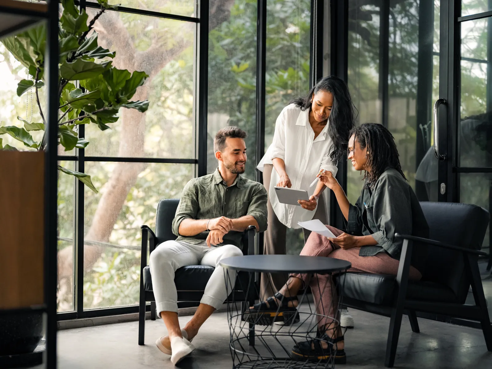 Three colleagues discuss documents and digital tablet in a modern office with large windows and plants.