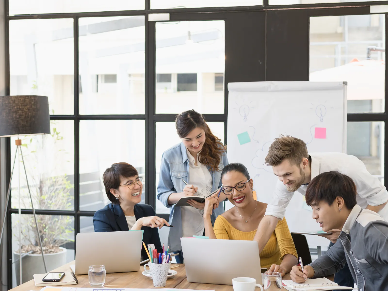 Team of five diverse young professionals collaborating around a table with laptops and notes in a modern office.
