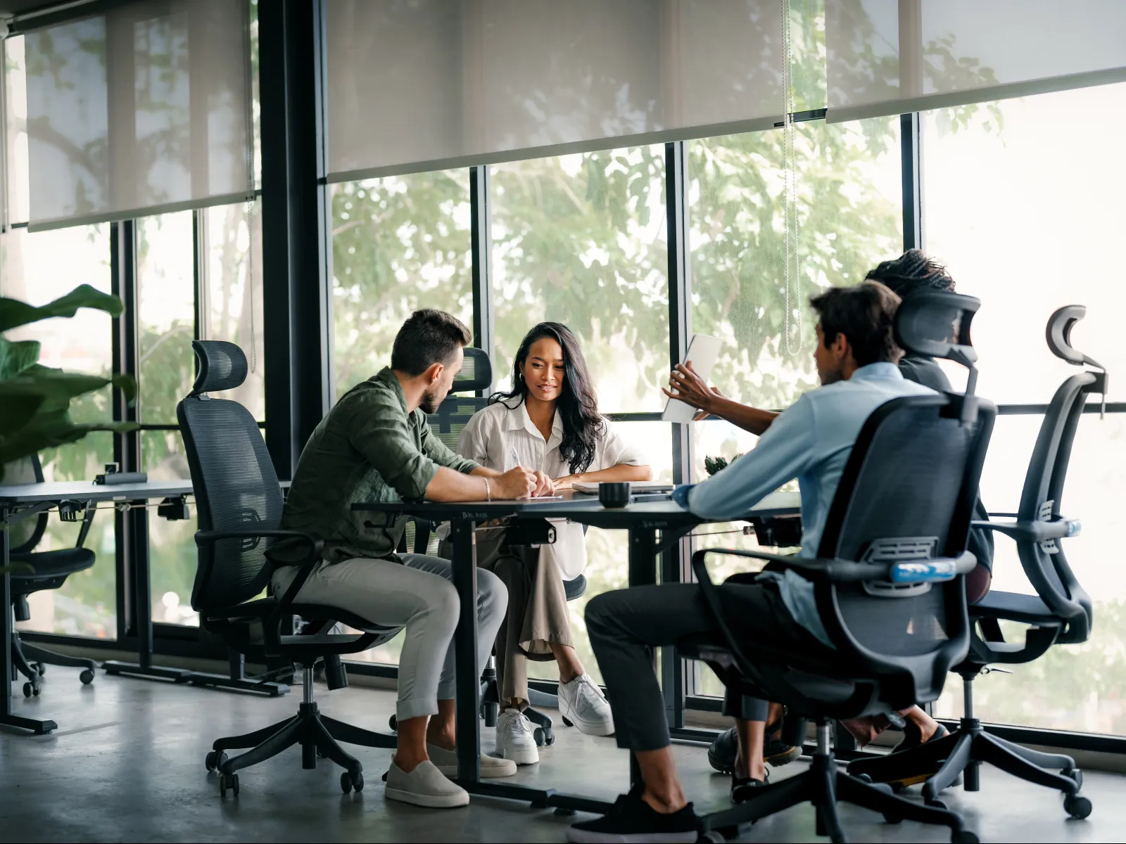 Four colleagues engaged in a discussion around a table in a modern office with large windows and natural light.