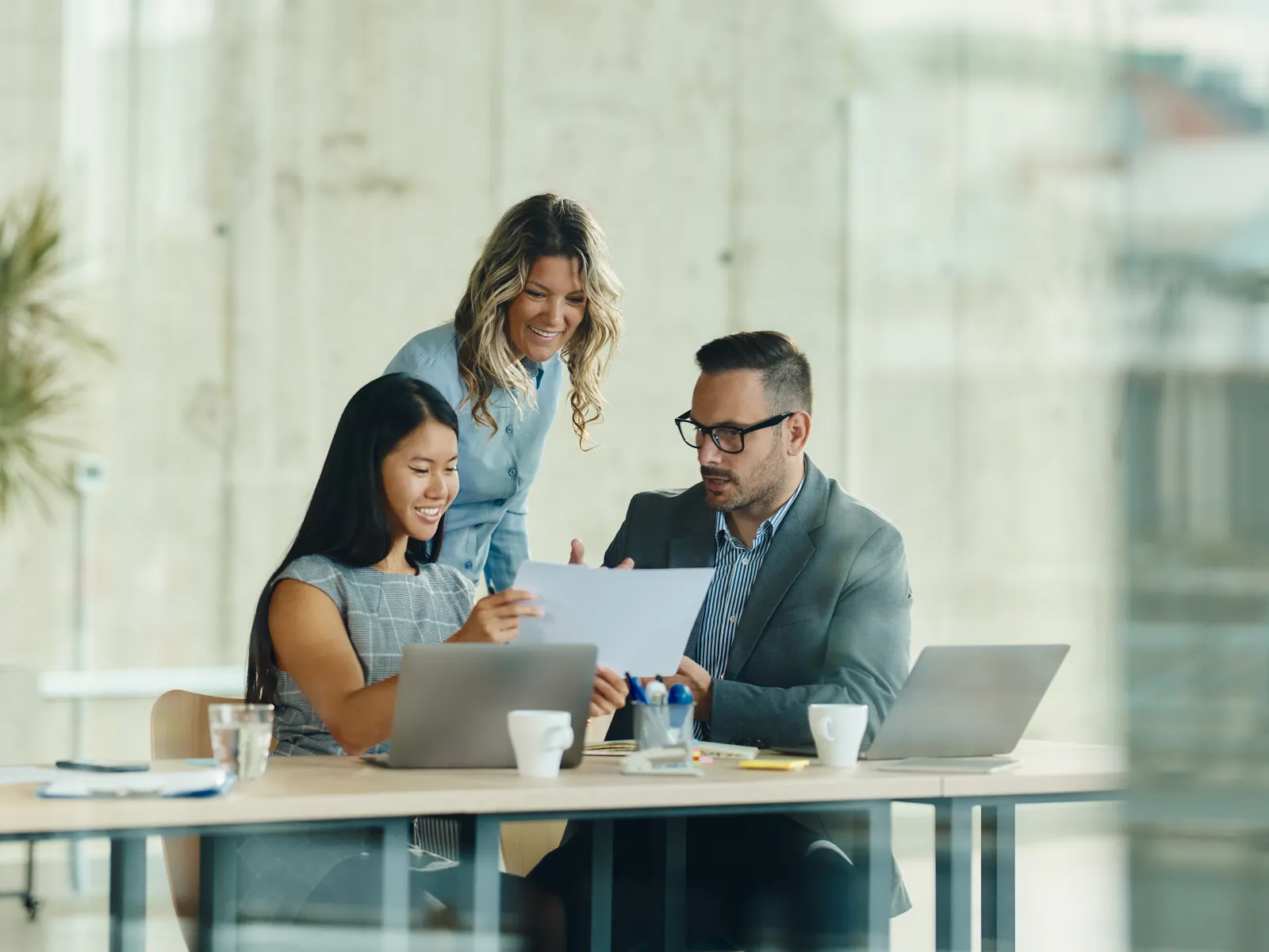 Three professionals reviewing documents and working on laptops in a modern office setting with large windows.