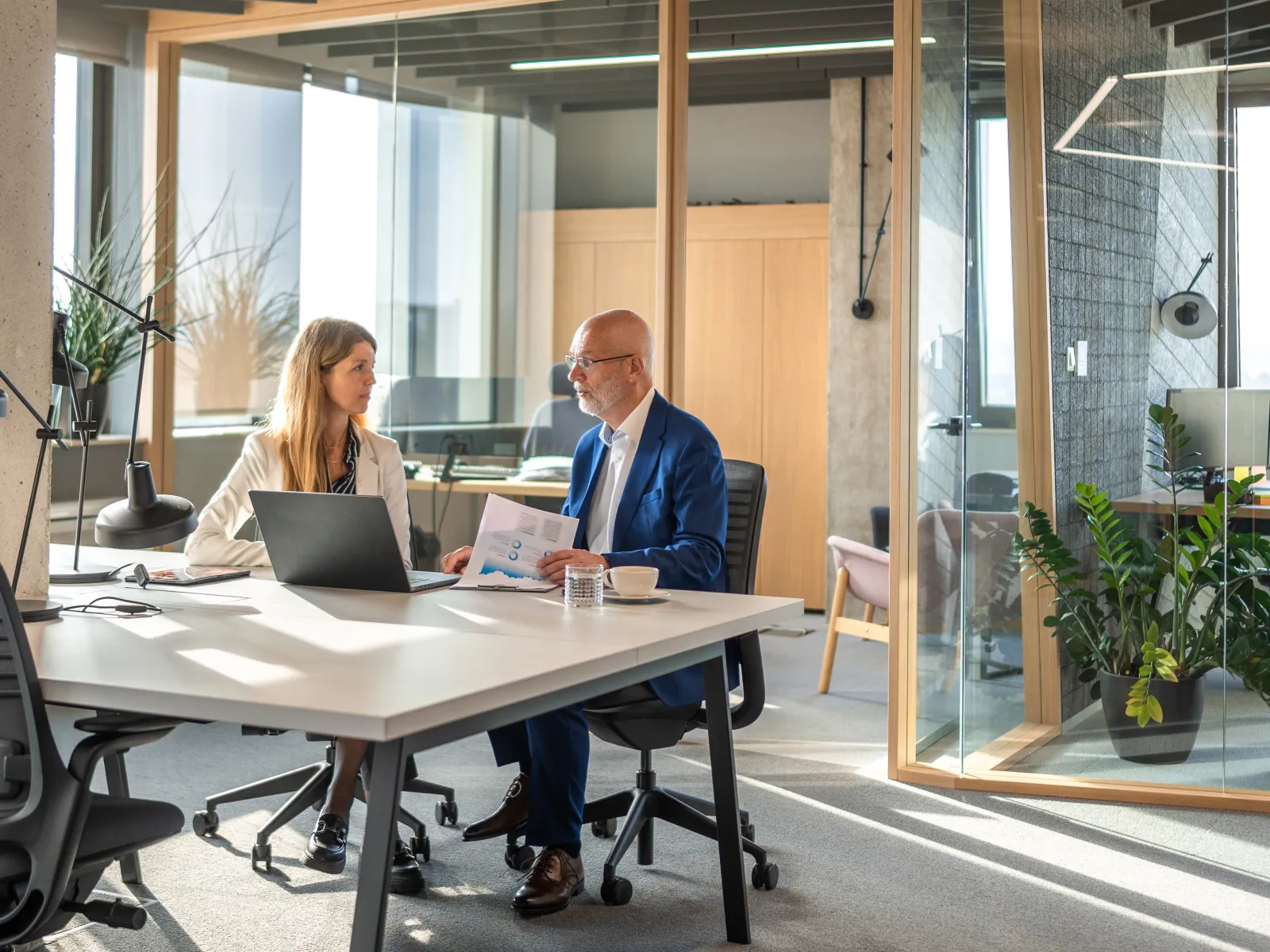 Two professionals discussing documents at a modern office table with laptop and glass walls.
