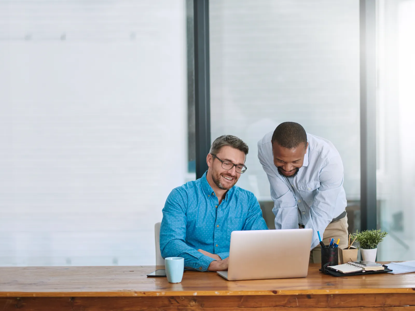 Two male colleagues collaborate smiling at a laptop in a bright modern office with coffee and stationery on the desk.