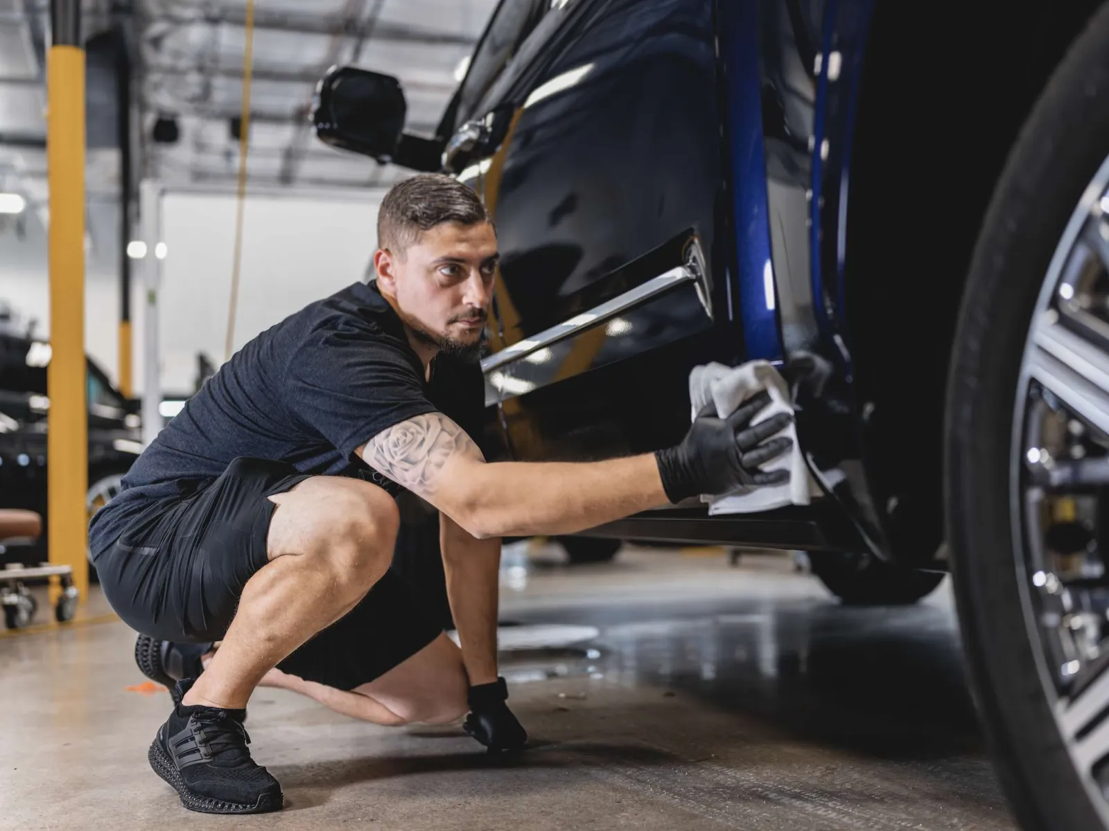 Man wearing black gloves carefully polishing a dark blue car in a well-lit garage.