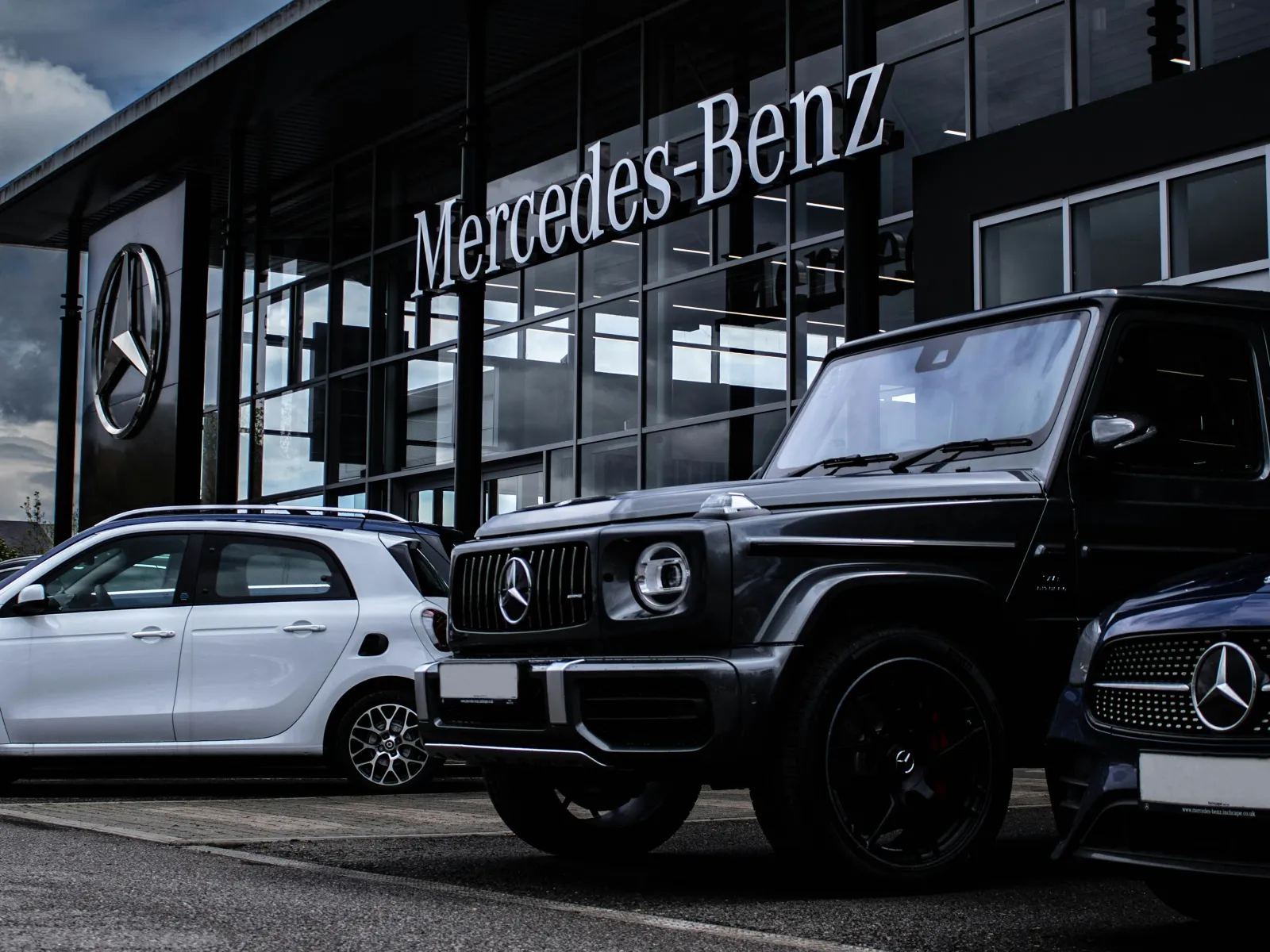 Mercedes-Benz dealership exterior with multiple luxury vehicles parked in front during cloudy day.
