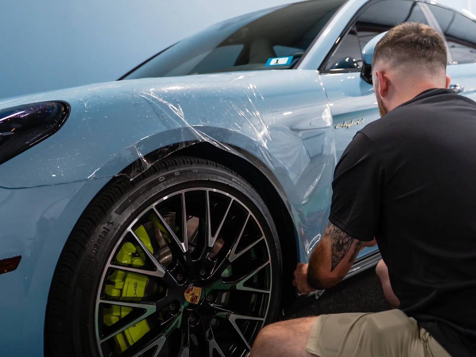 Man applying protective clear film wrap to blue Porsche sports car wheel arch and door.