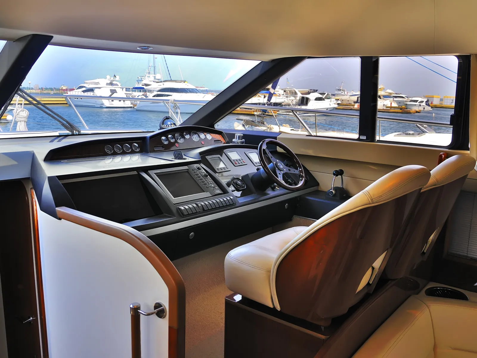 Interior view of a yacht cockpit with steering wheel, navigation instruments, and marina visible through windows.