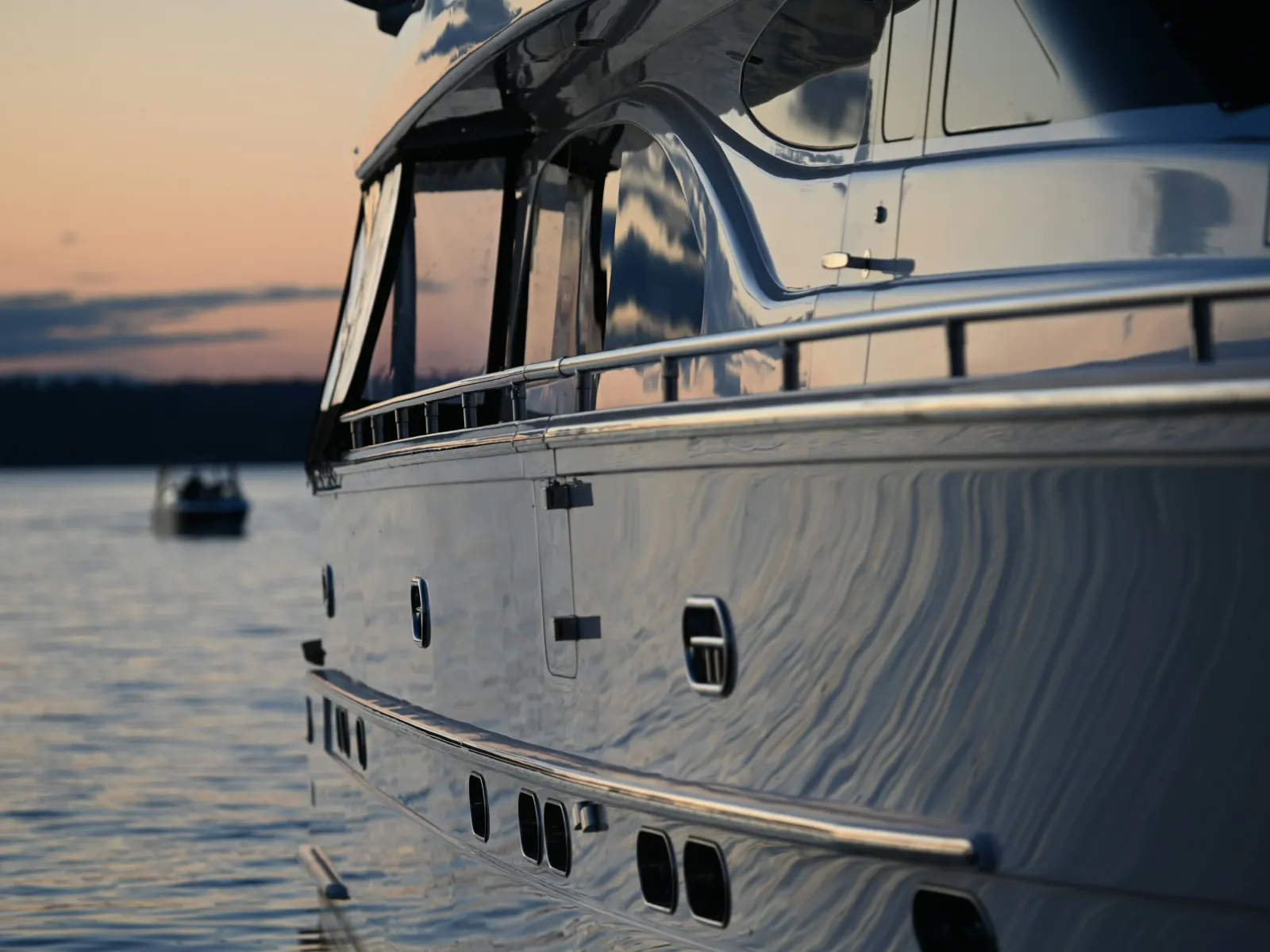Close-up of a yacht's sleek side reflecting the sunset over calm water with a distant boat in background