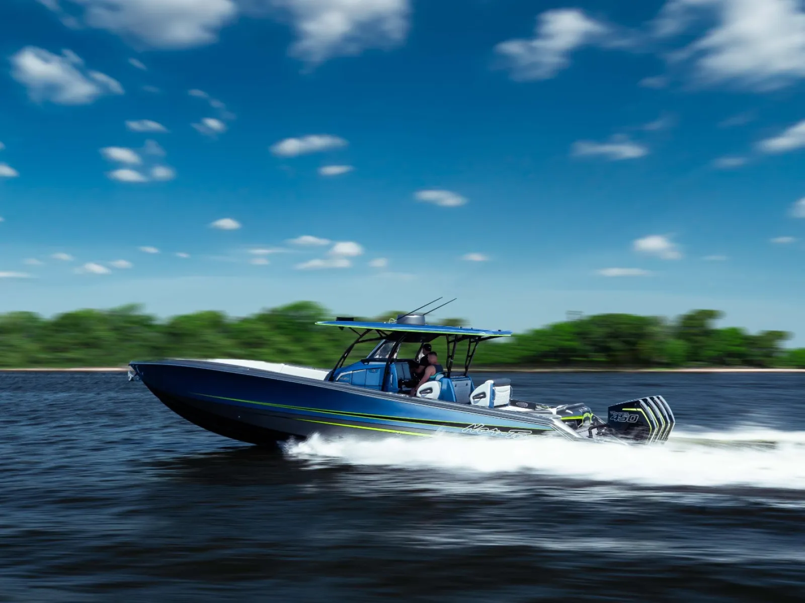 Speed boat cruising fast on water with blue sky and clouds overhead and green shoreline in the background.
