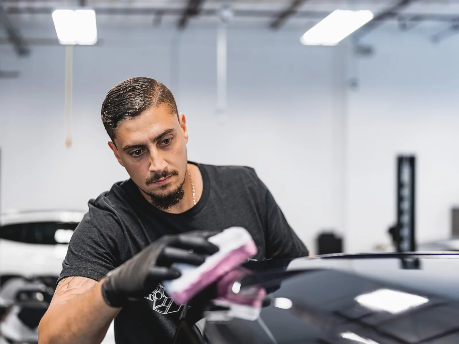 Man wearing black gloves polishing a black car hood in a bright auto workshop environment