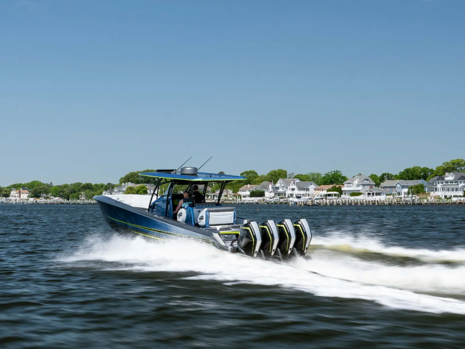 High-speed powerboat with four outboard engines cruising on water near a residential shoreline under clear blue sky.