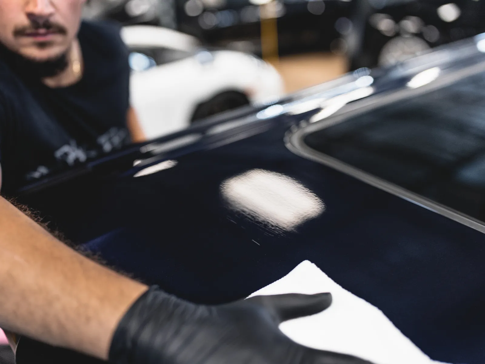Person wearing black gloves polishing a shiny black car surface with a white cloth inside a garage.