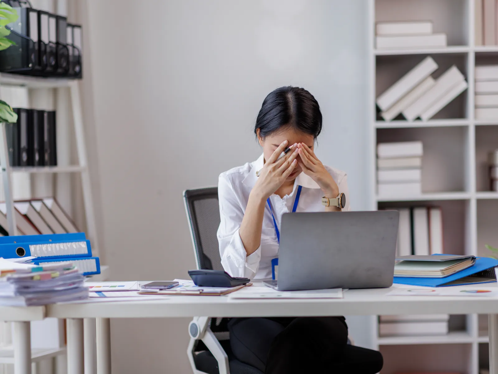 Overwhelmed woman in office stressed and rubbing her eyes sitting at desk with laptop and paperwork.