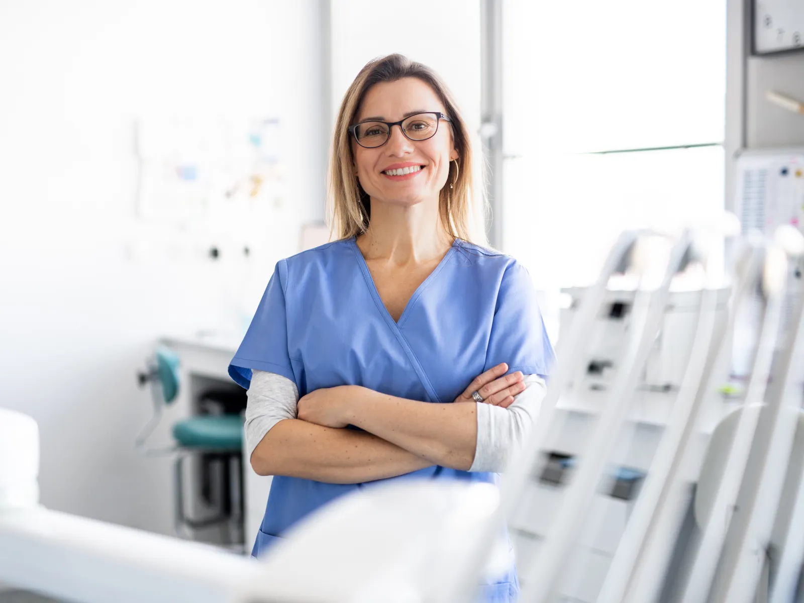 Smiling female dentist in blue scrubs standing confidently with arms crossed in a bright dental clinic.