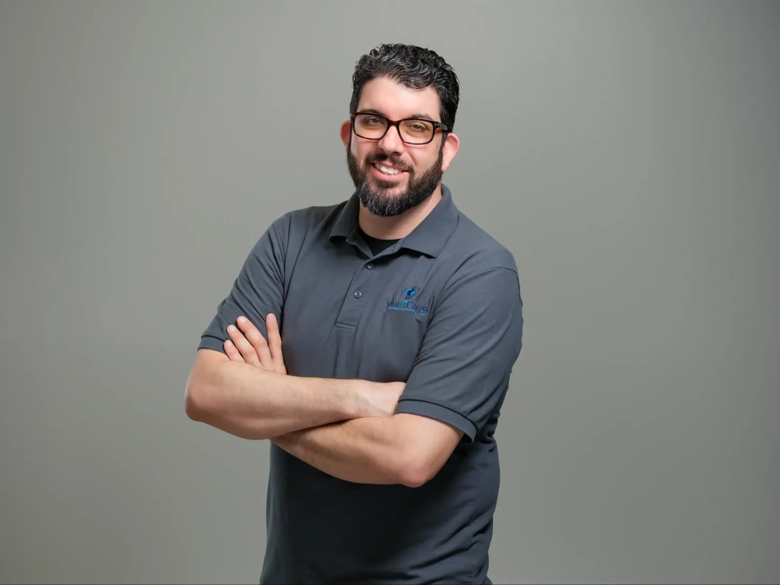Smiling man with glasses and beard wearing a gray polo shirt stands with arms crossed against a plain background.