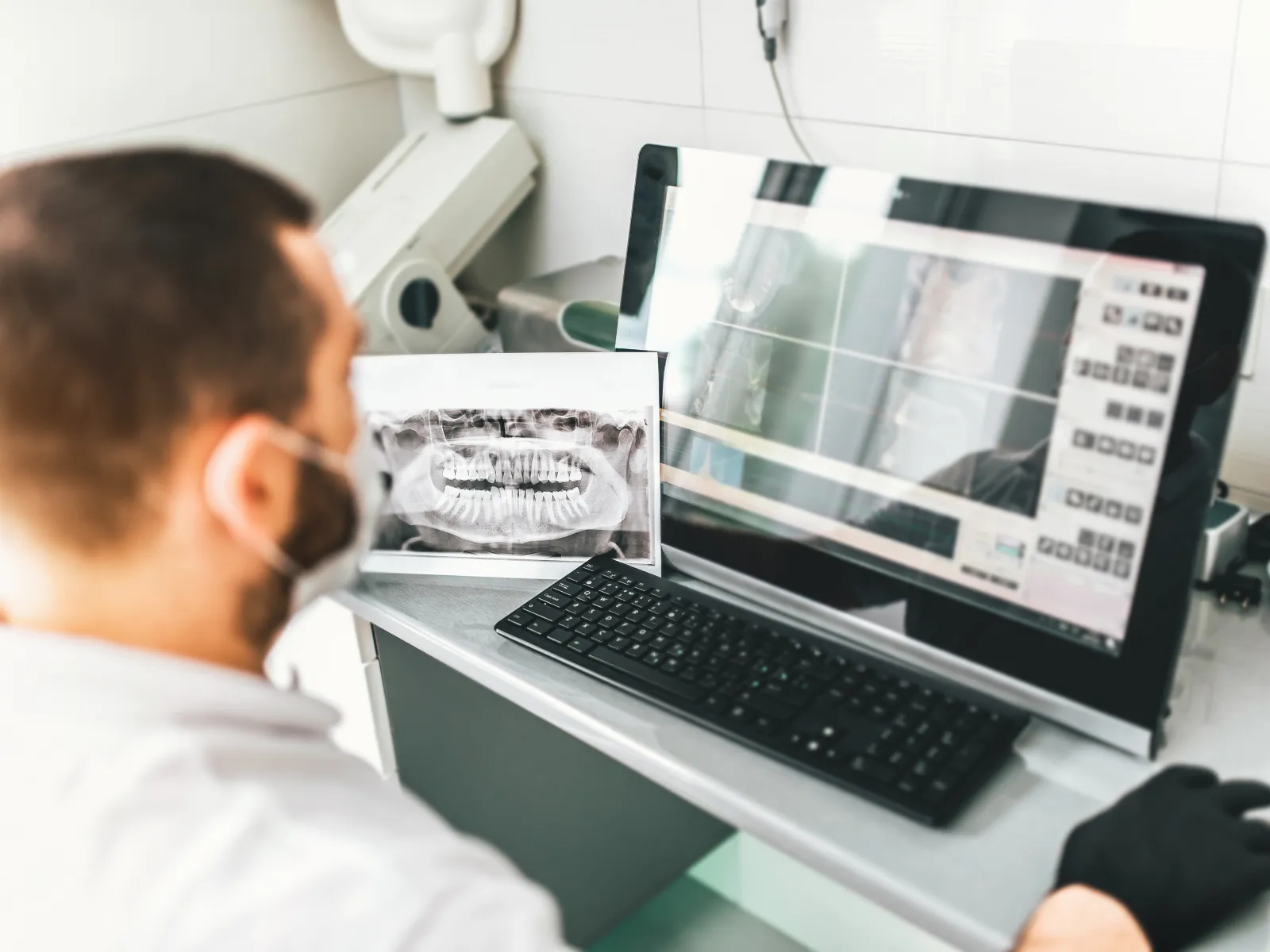 Dentist analyzing digital dental X-rays on computer screen with printed panoramic teeth X-ray nearby.