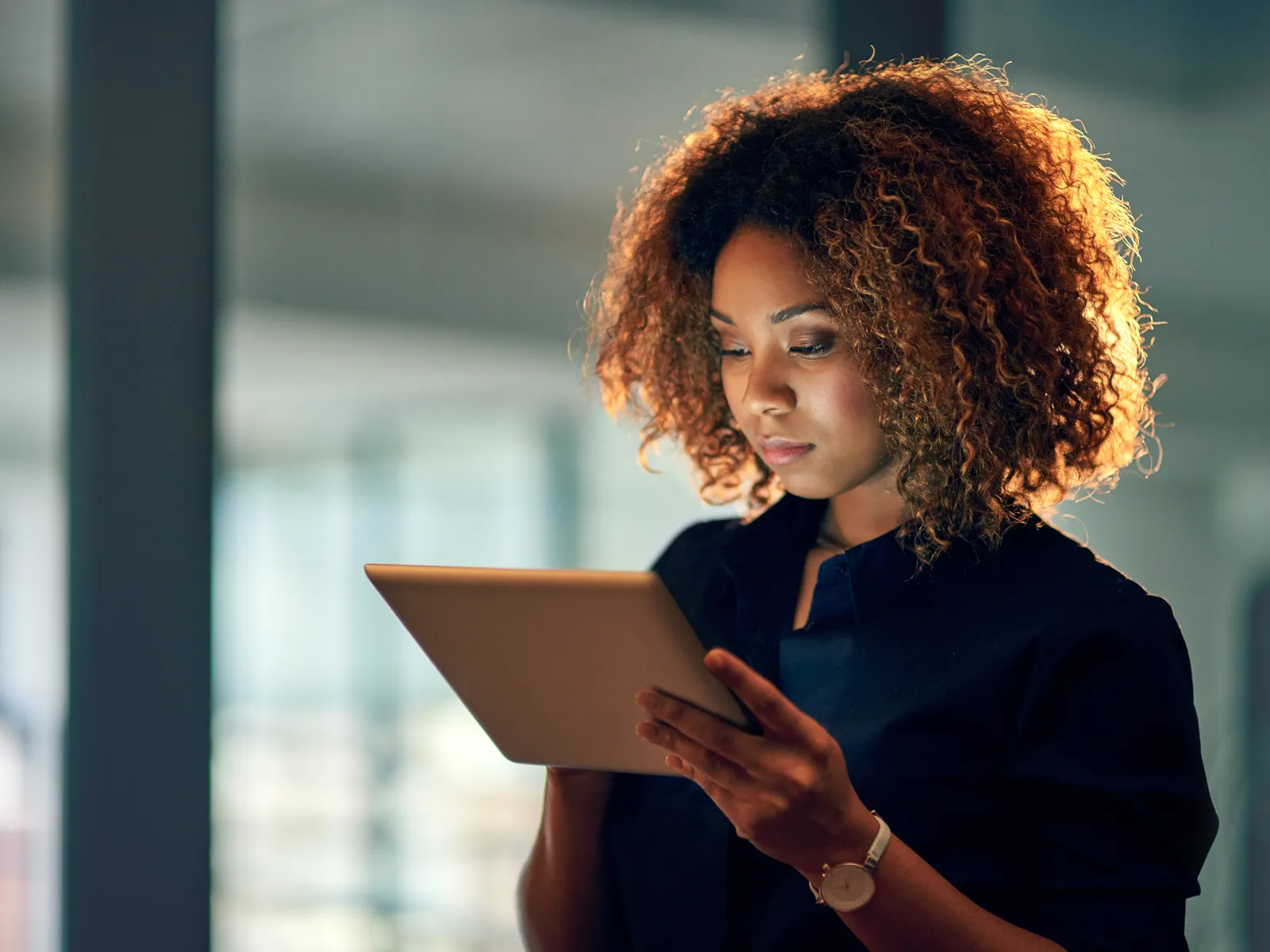 Young woman with curly hair using a digital tablet in a modern indoor setting, focused and engaged.