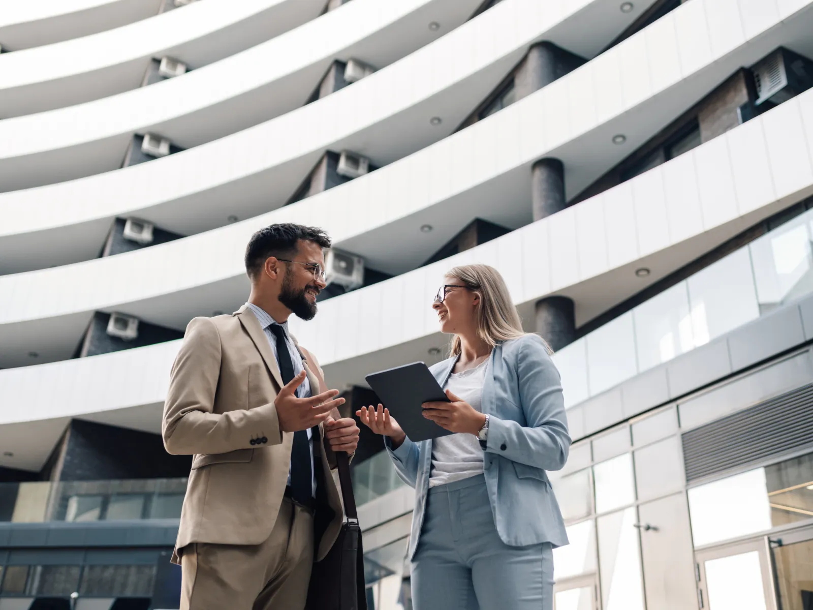Two business professionals, a man and woman, discussing work outside a modern office building with curved balconies.