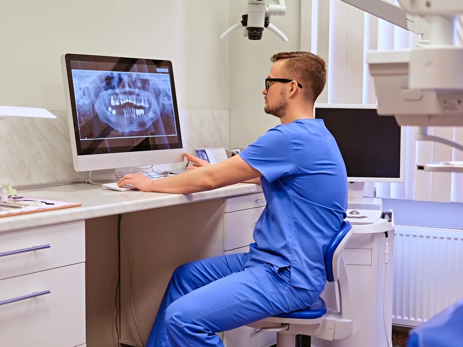 Dentist in blue scrubs reviewing dental X-ray on computer in modern clinic setting.