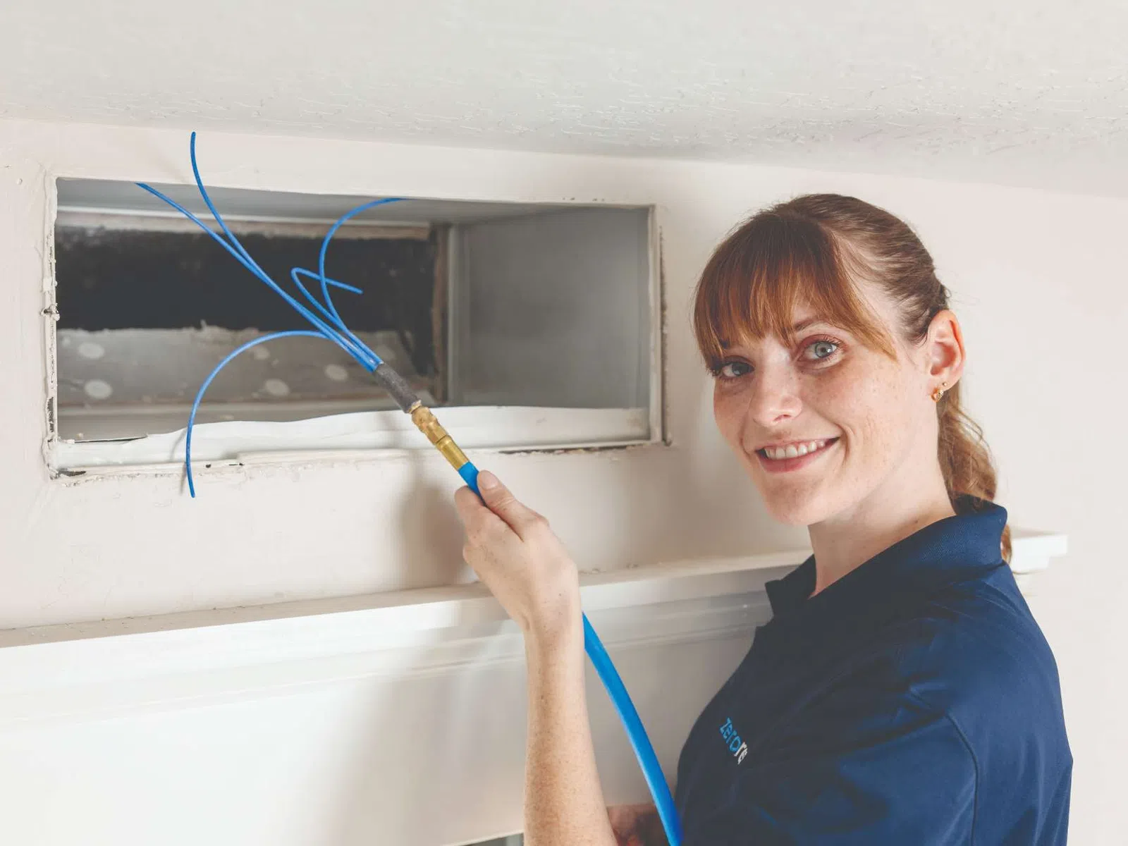 Technician in blue shirt using a flexible rod to clean ductwork inside a rectangular vent opening in the wall.