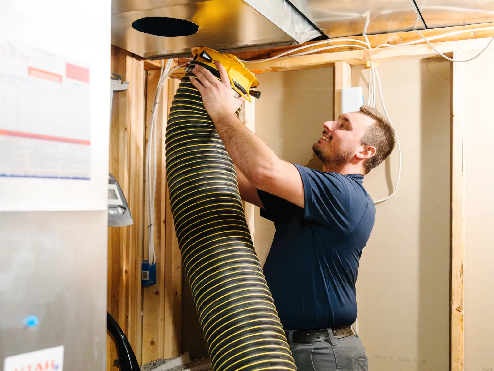 Technician installing flexible vent duct connected to HVAC system in unfinished basement with wooden framing.