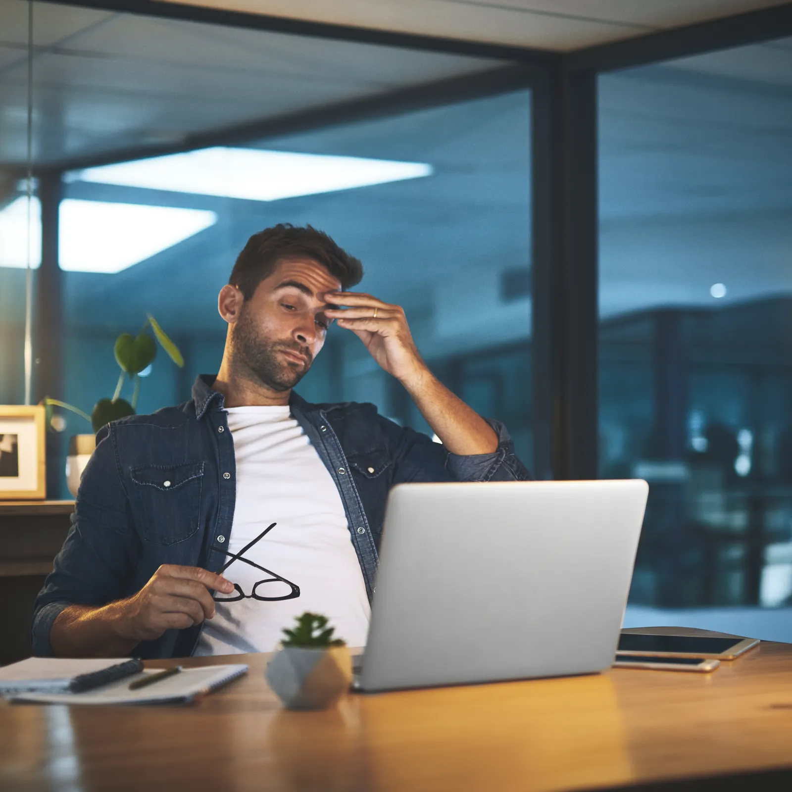 Man looking stressed while working late on a laptop in a modern office space.