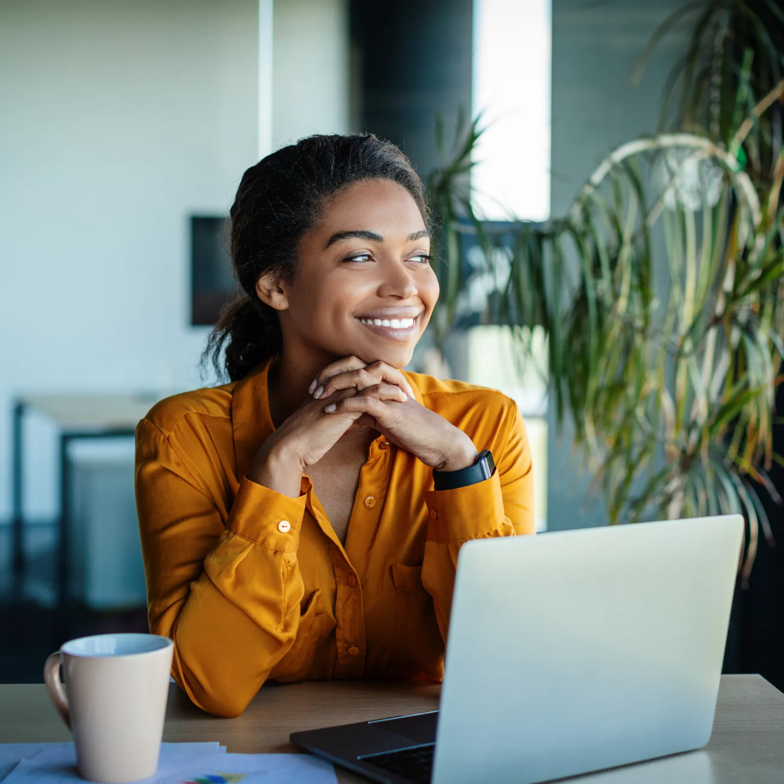 Smiling woman in a cozy office, looking at her laptop with a coffee cup nearby and greenery in the background.