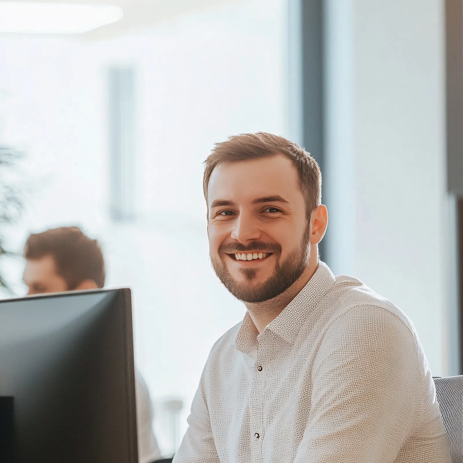 Smiling man in a modern office setting, seated at a desk with computer, exuding professionalism and positivity.