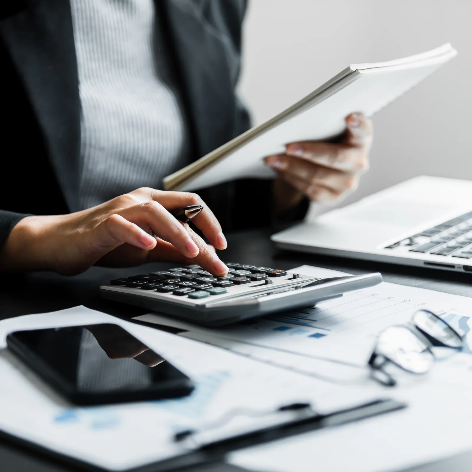 A person using a calculator while reviewing documents and working on a laptop at a desk.
