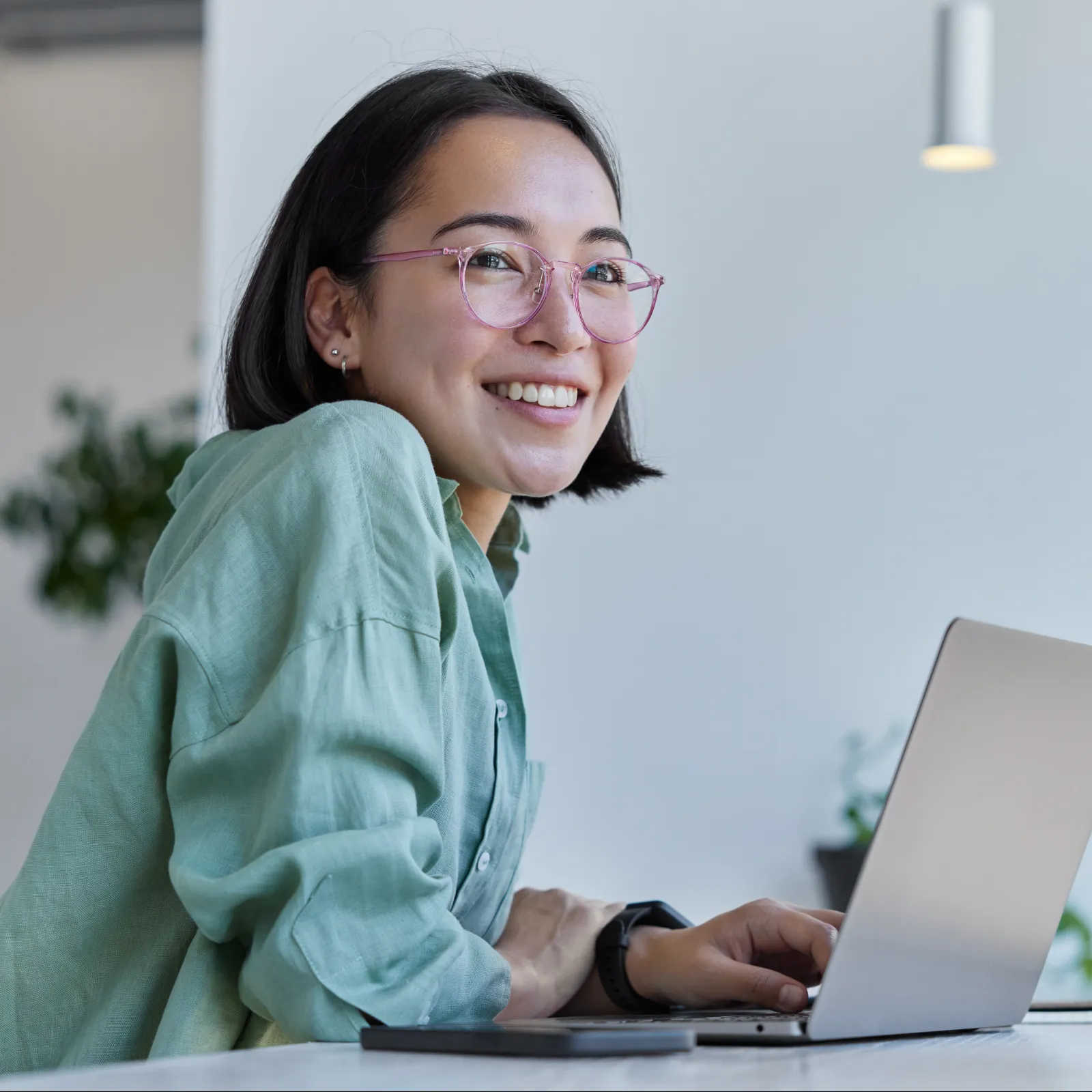 Smiling woman with glasses working on a laptop in a modern office space.