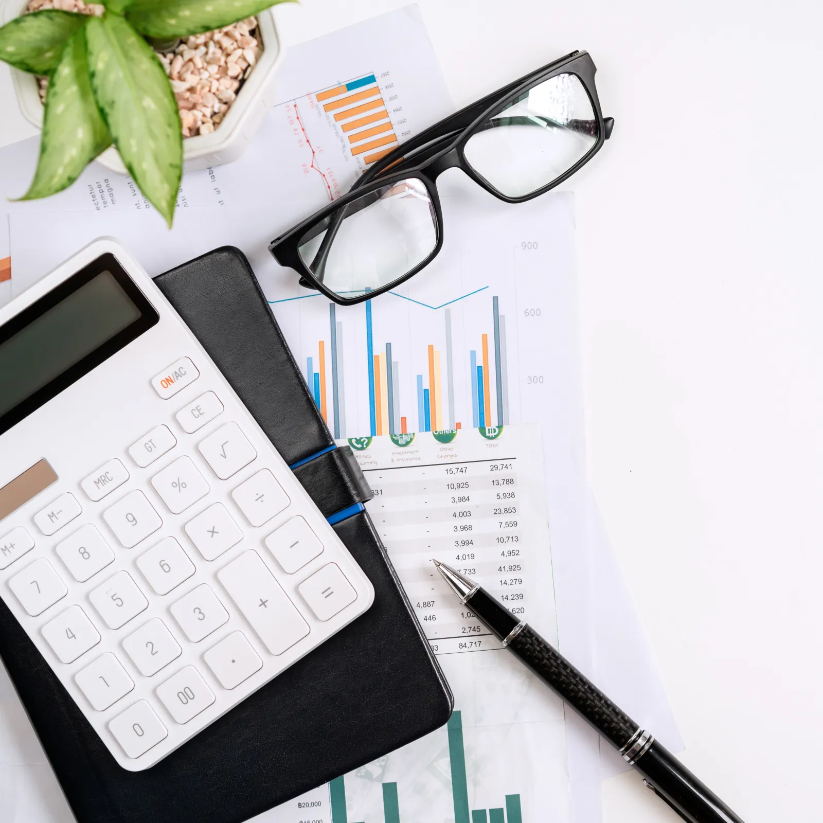 Flat lay of a calculator, glasses, notebook, and financial charts on a white desk.