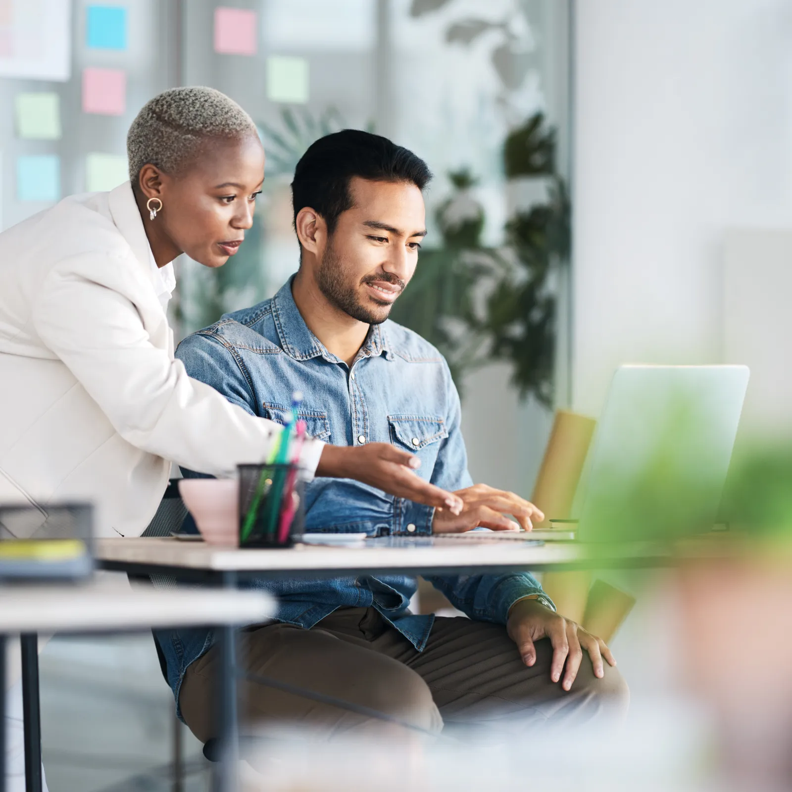 A businesswoman assists a man at a laptop in a modern office setting filled with colorful post-it notes.