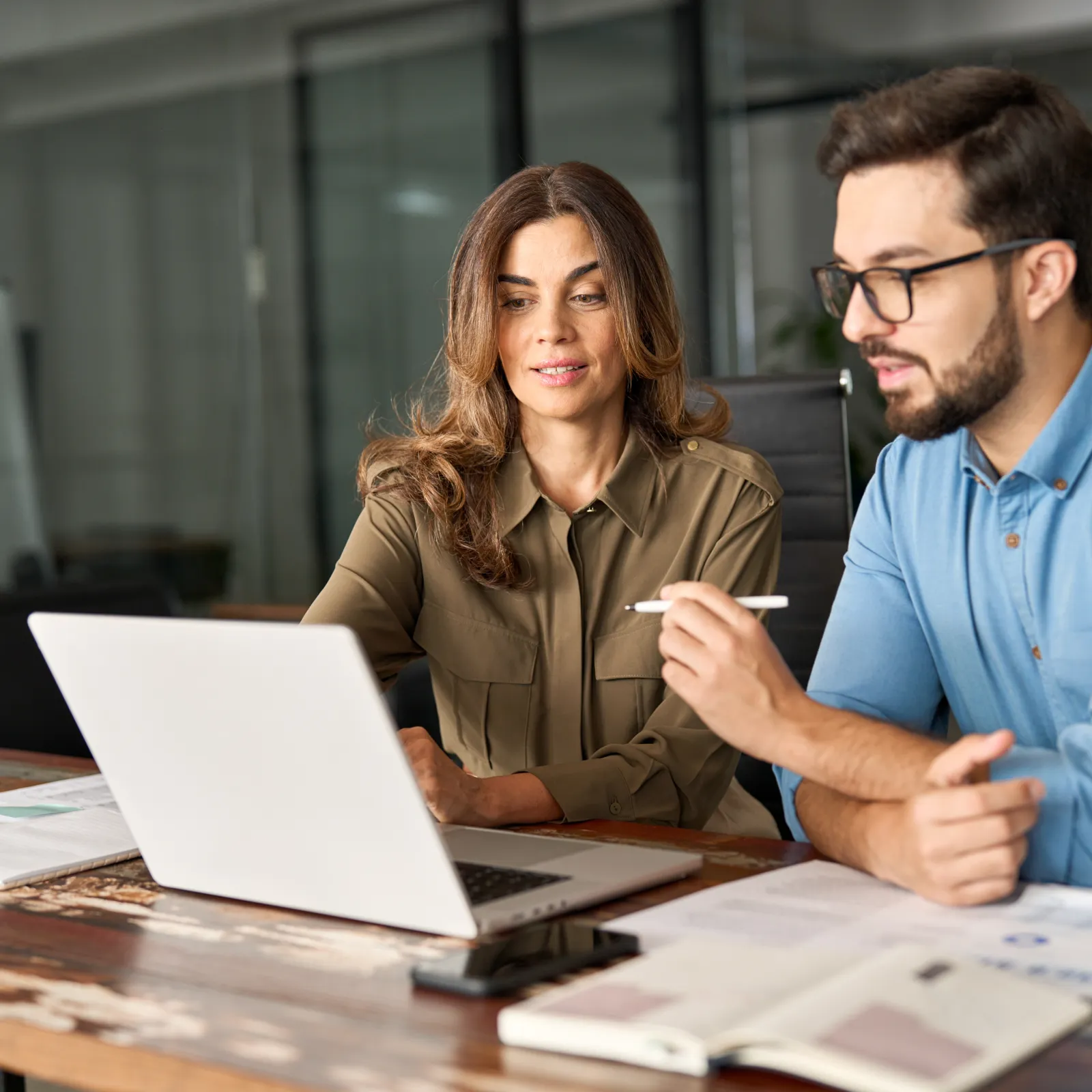 Two professionals collaborating in a modern office with a laptop and documents on a wooden table.
