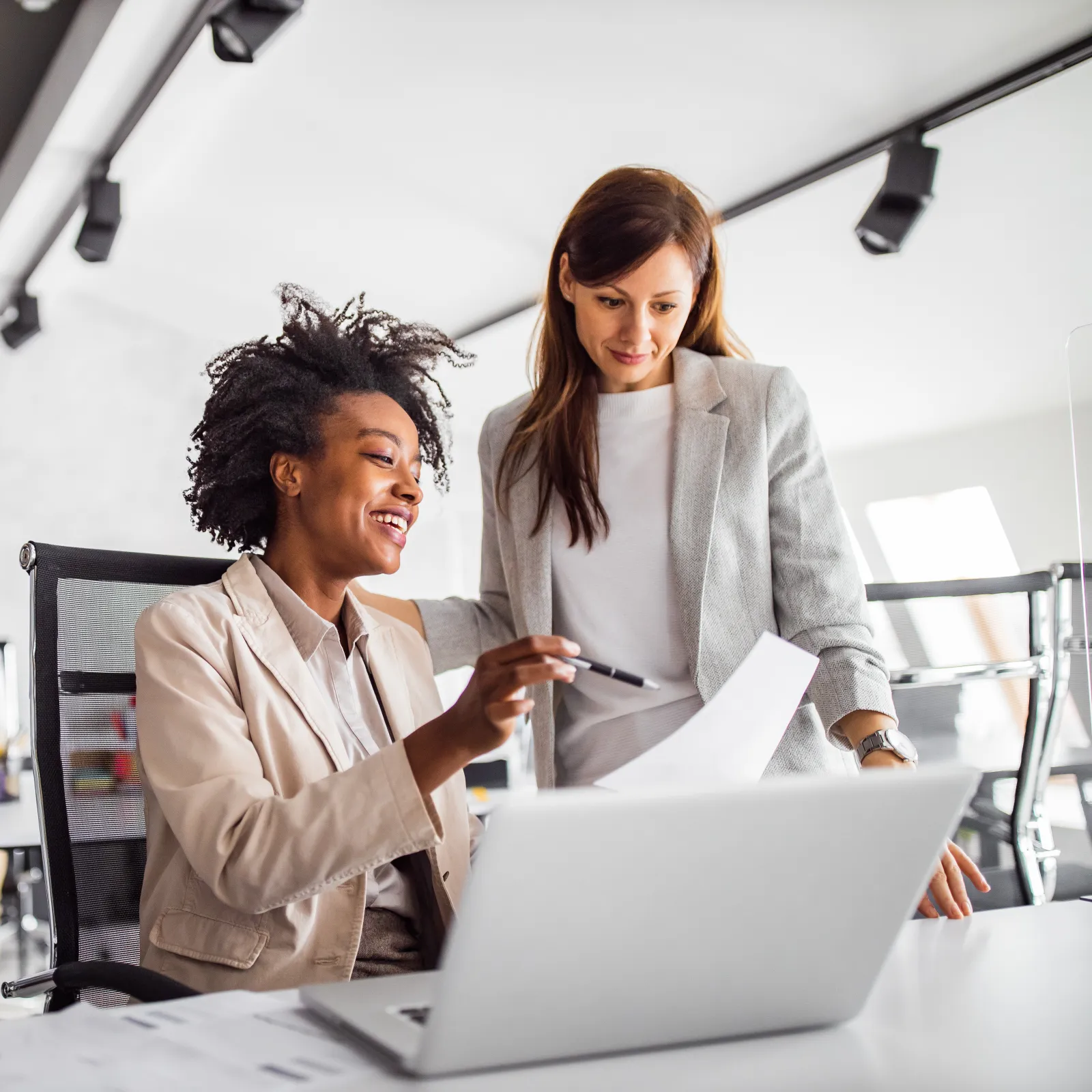 Two businesswomen collaborating in a modern office environment with a laptop and documents.