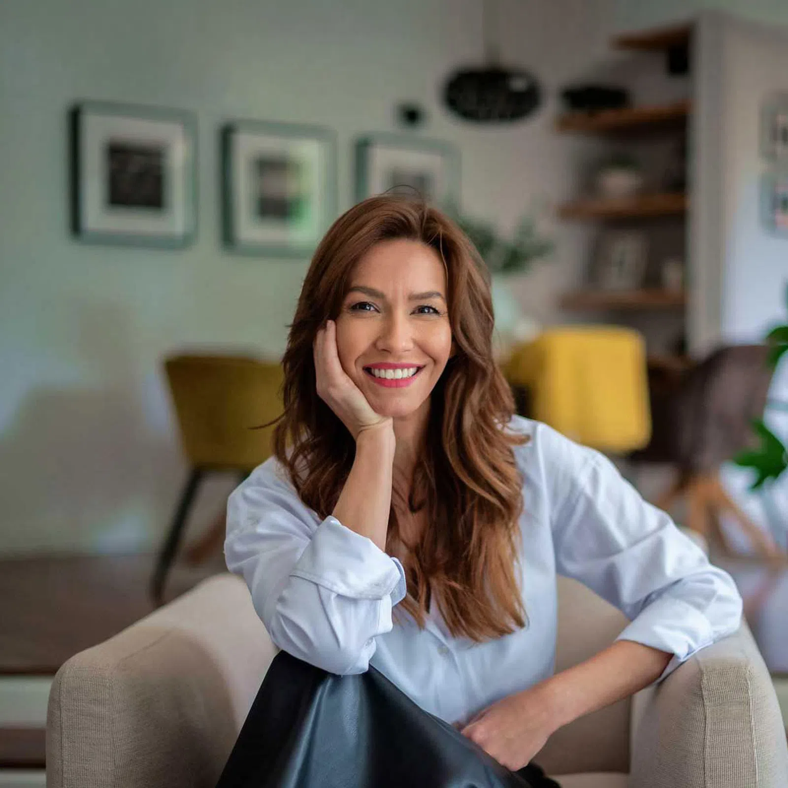 Smiling woman with long hair sitting on a beige armchair in a modern living room with plants and artwork.