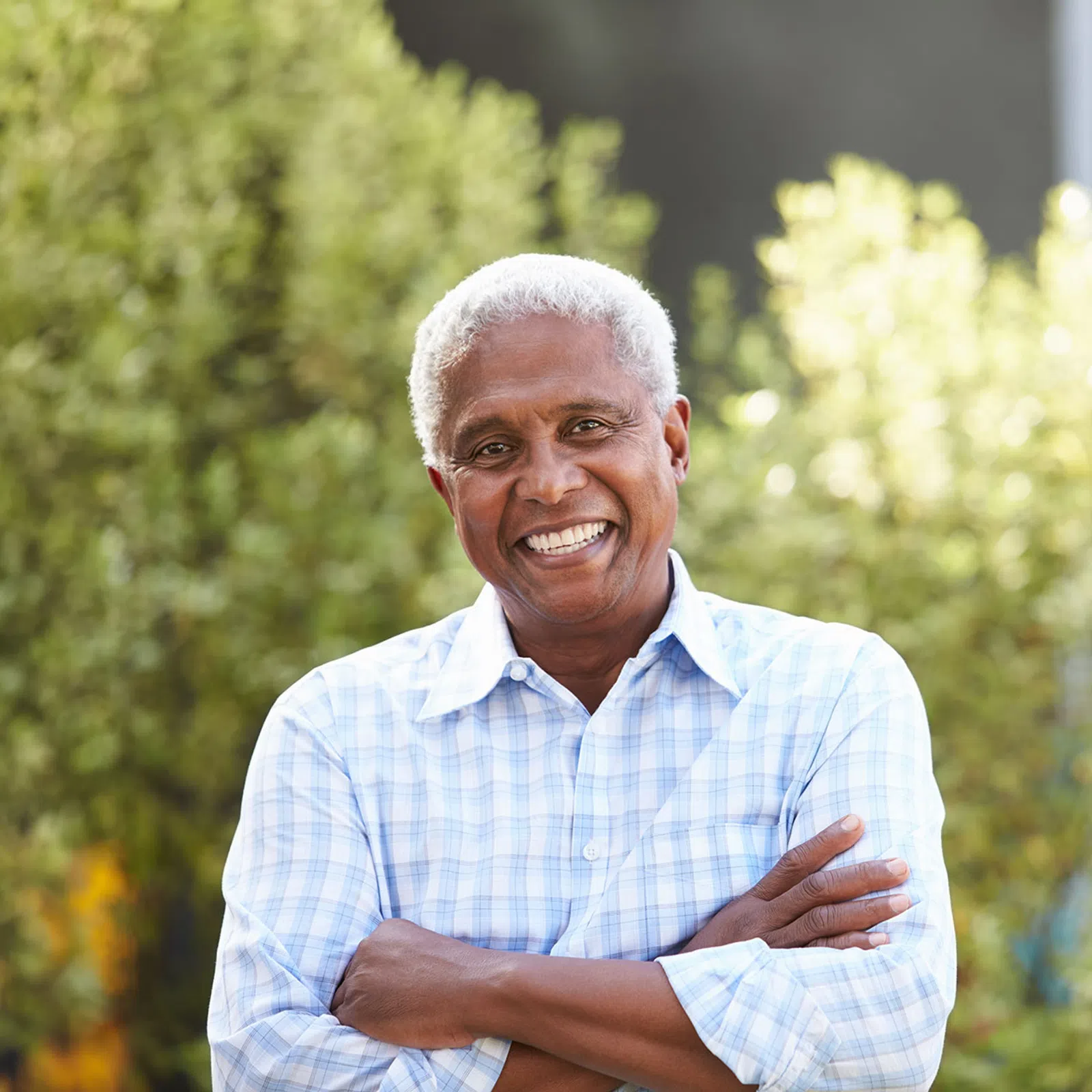 Smiling elderly man with gray hair in a blue checkered shirt standing outdoors with arms crossed, greenery background